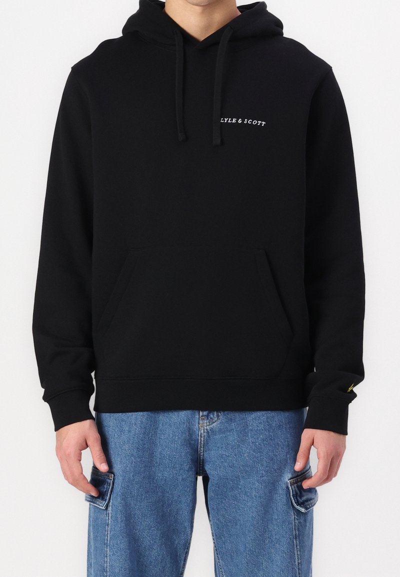 Homme portant un hoodie noir Lyle & Scott avec poche avant et un jean cargo bleu, se tenant contre un fond blanc uni.