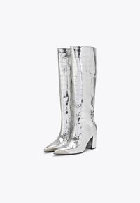 Błyszczące, srebrne botki do połowy łydki o teksturze krokodyla, ze spiczastymi czubkami i blokowymi obcasami. Gładkie metaliczne wykończenie, kanciasty design.