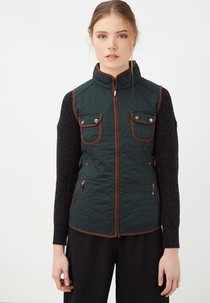 Chaleco acolchado verde con detalles marrones, que cuenta con un alto collar, dos bolsillos en el pecho y bolsillos laterales con cremallera. Llevado sobre una blusa negra texturizada.