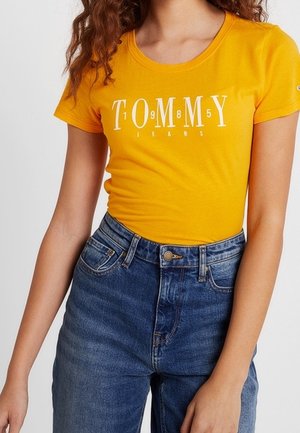 Kvinde iført en tætsiddende gul Tommy Jeans t-shirt med hvid logo tekst og højtaljede blå denim jeans, vist fra nakke til hofter.