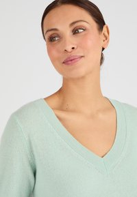 Pull léger vert menthe en matériau tricot doux, présentant un design à col en V et des accents côtelés au niveau du col et des poignets.