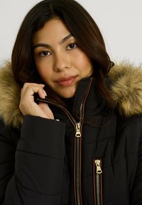 Jeune femme portant une doudoune noire avec une capuche bordée de fourrure, tenant le col près du visage, fond neutre.