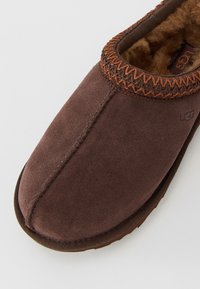 Brown suede slip-on slipper with fuzzy interior lining and decorative stitching around the opening, UGG logo visible on side and insole.