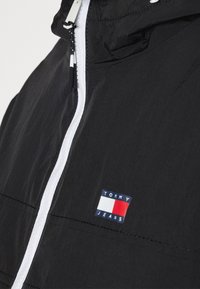 Zwarte windbreaker jas met een witte rits, verstelbare capuchon en een rechthoekige logo patch met blauwe, witte en rode kleurblokken.