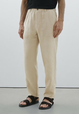 Beige Leinenhose mit lockerer Passform, ausgestattet mit zwei Fronttaschen und einem Knopfverschluss. Kombiniert mit schwarzen Riemchensandalen.