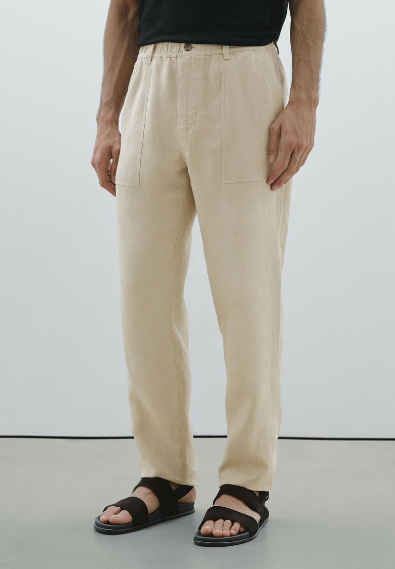 Pantalon en lin beige avec une coupe décontractée, comprenant deux poches avant et une fermeture à bouton. Associé à des sandales noires à lanières.