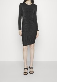 Svart långärmad klänning med rynkade detaljer och en figurnära silhuett. Glänsande tyg ger textur. Bärs med svarta, remmar sandaler.