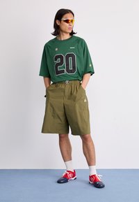Green mesh jersey with large black "20," olive shorts with elastic waistband, red and white shoes, and reflective yellow sunglasses.