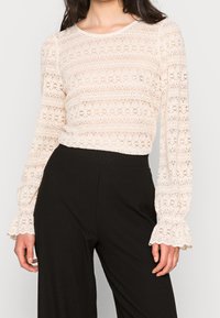 Haut en dentelle à manches longues de couleur beige clair, avec un motif floral, un col rond et des poignets évasés. Associé à un pantalon large à côtes noir.