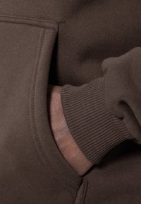 Sudadera marrón con un bolsillo lateral y puños acanalados. La tela parece suave y texturizada, con detalles de costura visibles y bien hechos.
