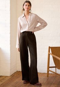 Chemise à manches longues rayée beige et blanche, associée à un pantalon large taille haute noir et des chaussures marron épaisses. Sol en bois en arrière-plan.