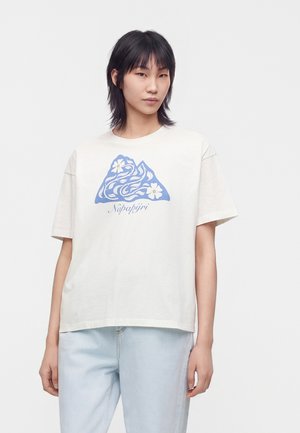 Jeune personne aux cheveux noirs et raides portant un t-shirt blanc avec un motif floral bleu montagne et un jean bleu clair.