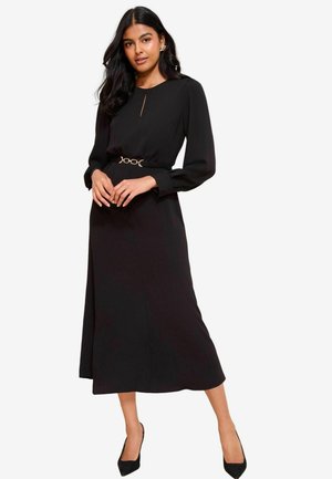 Langes schwarzes Kleid mit langen Ärmeln, einem runden Halsausschnitt, einem Schlüssellochdetail und einer taillierten Taille, die durch einen Gürtel in Goldoptik betont wird.