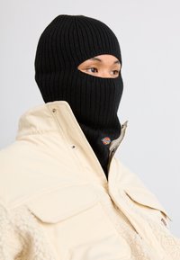Sort strikket balaclava med ribbet tekstur, båret under en cremefarvet polstret jakke, der har to lommer og et logo-patch.