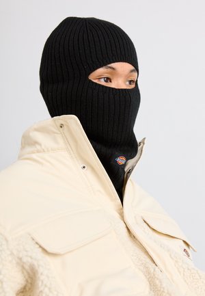 Sort strikket balaclava med ribbet tekstur, båret under en cremefarvet polstret jakke, der har to lommer og et logo-patch.