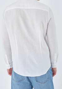 Camicia bianca di lino con maniche lunghe, polsini botttonati e orlo curvo. Texture morbida e aspetto leggero. Vista posteriore in evidenza.