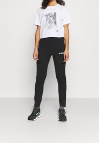 Pantalones cargo negros con bolsillos con cremallera y detalles de logo, combinados con una camiseta blanca corta que presenta un estampado gráfico en tonos de gris.