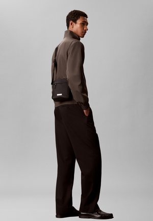 Joven de pie de lado con chaqueta marrón, pantalones negros, mocasines y un pequeño bolso negro cruzado sobre un fondo gris.