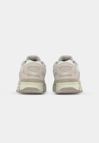 Pár béžových a bielych tenisiek Reebok zobrazených zozadu, zdôrazňujúcich polstrovanú podrážku a logo značky na päte.
