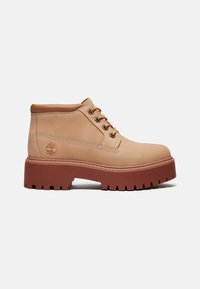 Bottine en cuir tabac avec col matelassé marron, semelle épaisse rouge-brun à crampons, logo latéral et fermeture à lacets sur fond blanc.