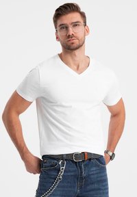T-shirt bianco a scollo a V in cotone, indossato con jeans blu, una cintura a fantasia e un dettaglio in catena argentata. Occhiali trasparenti su un modello maschile.
