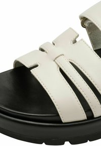 Sandal in pelle bianca con tre cinturini, suola nera testurizzata e accentuazione in metallo circolare. Caratterizzata da un design piatto con una finitura liscia.