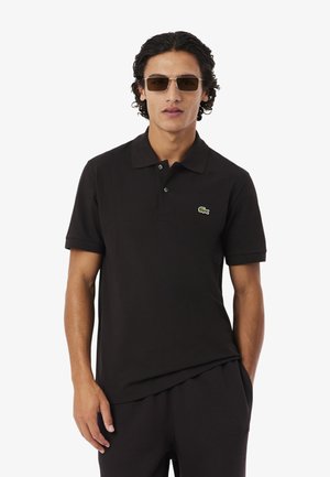 Homme portant un polo à manches courtes noir avec un logo crocodile vert et des lunettes de soleil rectangulaires marron, debout avec une main dans la poche.