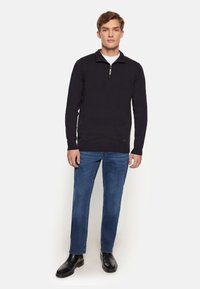 Pulover din tricot cu guler la fermoar în nuanța marină, asortat cu blugi albaștri cu croială dreaptă și botine negre cu gleznă. Design simplu cu o textură uniformă.