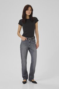 Naine seisab halli tausta ees, üks käsi taskus, seljas must lõikega t-särk, tuhmunud hallid teksad vööga ning mustad teravate ninadega kingad.