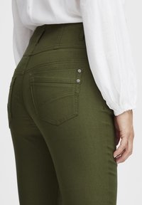 Jeans de tiro alto en verde oliva con bolsillos, que presentan una textura suave, detalles de costura sutiles y una silueta ajustada.