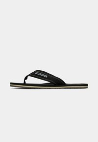 Tongs noires avec une semelle en caoutchouc noir texturé, dotées d'une sangle en tissu avec des coutures jaunes et d'un logo "HILFIGER" en blanc.