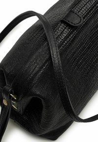 Černá texturovaná kabelka s hadím vzorem, se štíhlým černým popruhem a malým klopovým detailem poblíž horního zavírání.