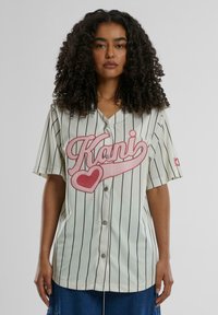 Kremfarget baseball-skjorte med svarte pinstriper, med stor rosa grafikktekst "Kani" og hjerte-design, laget av lett stoff.