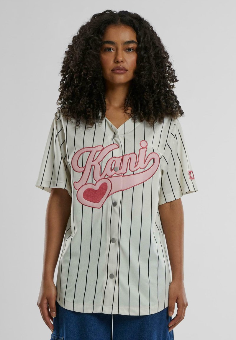 Kremfarget baseball-skjorte med svarte pinstriper, med stor rosa grafikktekst "Kani" og hjerte-design, laget av lett stoff.