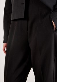 Close-up van een persoon in zwarte op maat gemaakte broek en bijpassende jacket, met één hand in de zak en de andere ontspannen naast hun lichaam.