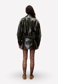 Veste en cuir noire surdimensionnée avec une taille froncée, associée à un collant imprimé léopard marron et des chaussons beige, vue de dos.