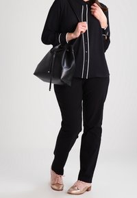 Femme en tenue noire tenant un sac à main en cuir noir, portant un pantalon noir et des chaussures à lacets métalliques couleur or rose, debout devant un fond uni.