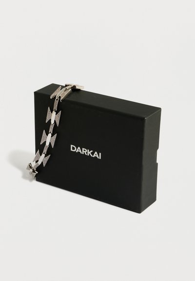 Srebrna zapestnica v obliki pentlje, prikazana poleg črne pravokotne škatle z belim napisom blagovne znamke "DARKAI".