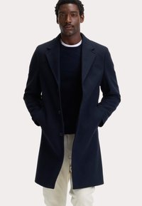 Marineblauwe jas van een wolmengsel met puntige revers, knopen aan de voorkant en steekzakken, gedragen over een marineblauwe trui en gecombineerd met lichtgekleurde broek.