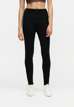 Femme portant un legging noir taille haute et des baskets blanches, debout devant un fond blanc uni.