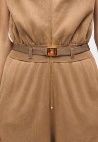 Φόρμα σε ταμπά χρώμα με φερμουάρ μπροστά, ελαστική μέση και καφέ υφασμάτινη ζώνη με χρυσή αγκράφα. Μαλακό, υφασμάτινο υλικό με υφή.