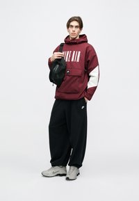 Casaco corta-vento bordo com o texto "NIKE AIR", mala de tiracolo preta, calças largas pretas e ténis cinzas com detalhes. Design minimalista.