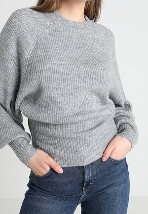 Donna che indossa un maglione a coste grigio chiaro con maniche lunghe e jeans blu, le mani infilate nelle tasche anteriori.