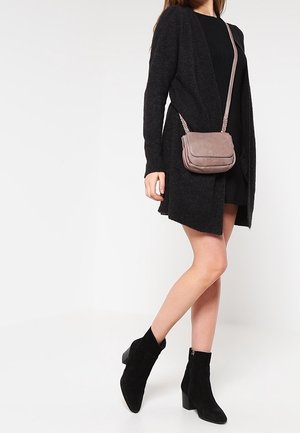 Mujer vistiendo vestido negro y cárdigan, botas negras hasta el tobillo y llevando un bolso bandolera color malva, de pie sobre un fondo blanco.