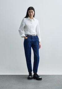 Camicia bianca con bottoni, pantaloni di jeans blu e scarpe nere stringate con punta lucida. Design semplice, vestibilità dritta, stile minimalista.