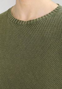 Maglione verde lavorato a maglia con un motivo testurizzato e scollo a costine. Il materiale sembra morbido con uno spessore medio.