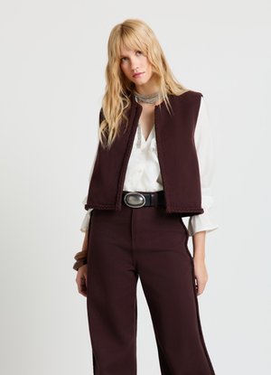 Bordeauxrotes Weste mit kastiger Form, aus dickem Stoff gefertigt, mit geflochtenem Besatz und ohne Verschlüsse. Kombiniert mit einer weißen Bluse und hoch taillierten Hosen.
