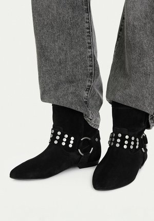 Czarne zamszowe botki za kostkę z srebrnymi paskami i pierścieniami z nitami, noszone z luźnymi szarymi dżinsami.