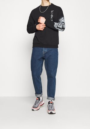 Person wearing black sweatshirt with white dragon sleeve print and Japanese text, blue rolled-up jeans, white and pink sneakers, and silver chain.