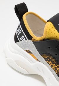 Baskets noires et jaunes avec une tige en mesh texturé, des accents pailletés argentés, et une semelle blanche avec un motif ondulé unique.